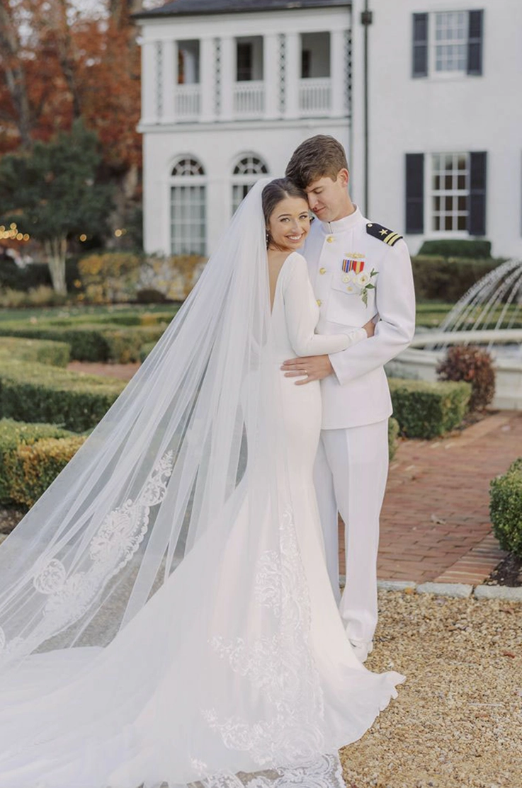 Groom embraces bride in a crepe fit and flare gown with a low back and flowing veil, in front of a fountain in an estate.
