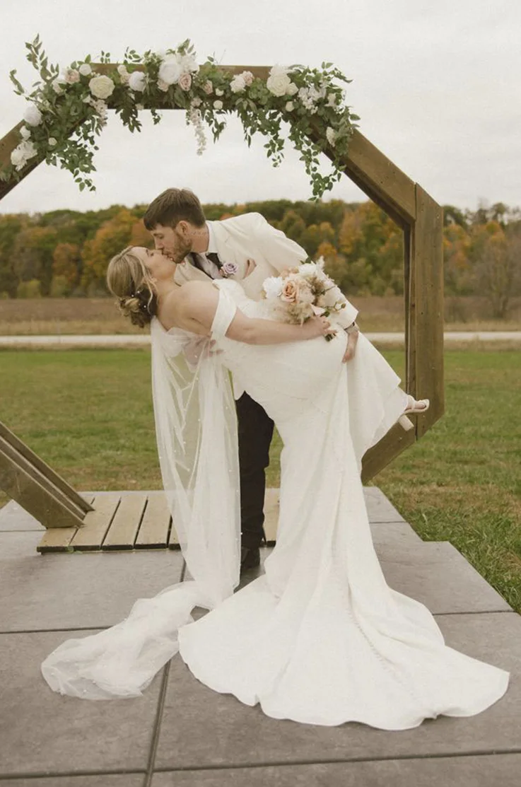 Groom dips his bride at a wooden altar near fall colored trees and in an off-shoulder crepe wedding dress, holding flowers. 