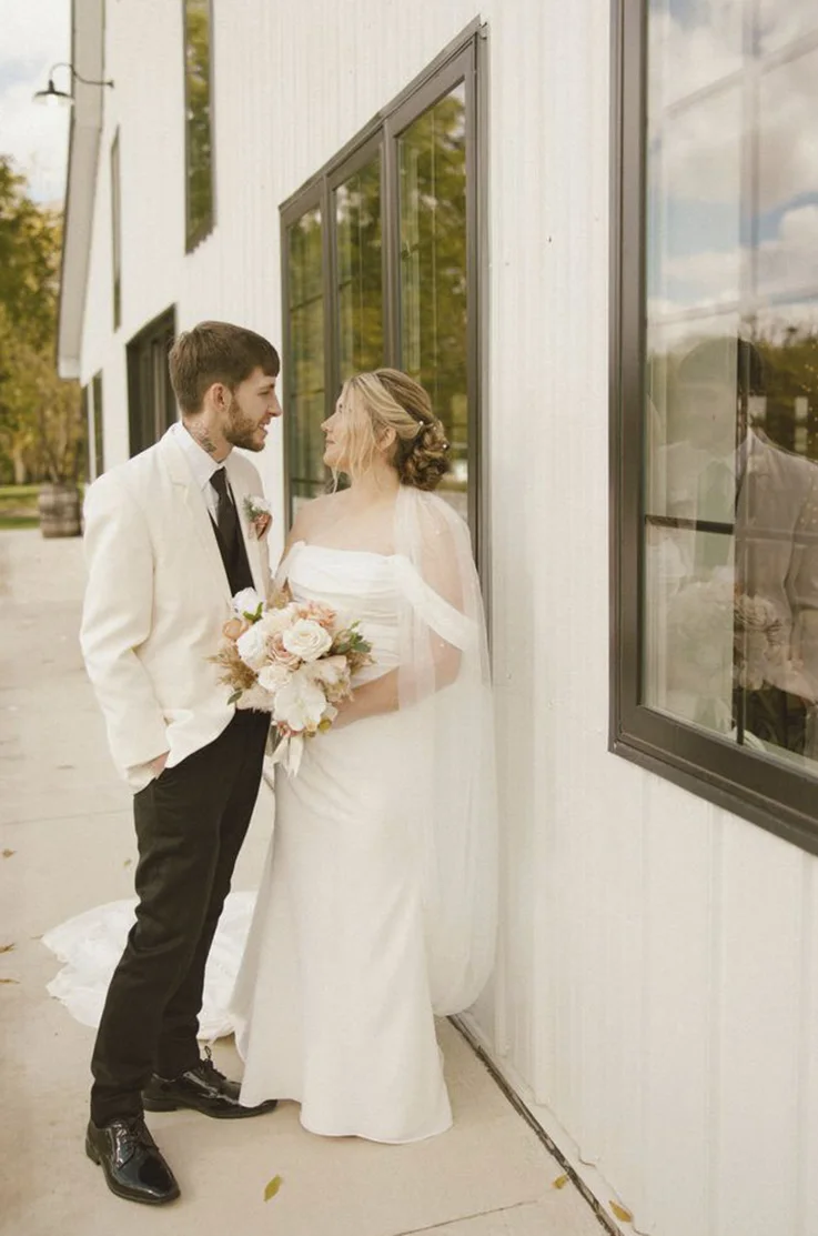 Bride in an off-shoulder crepe fitted gown with a veil and a scoop neckline, is holding a bouquet outside the venue and faces her groom. 