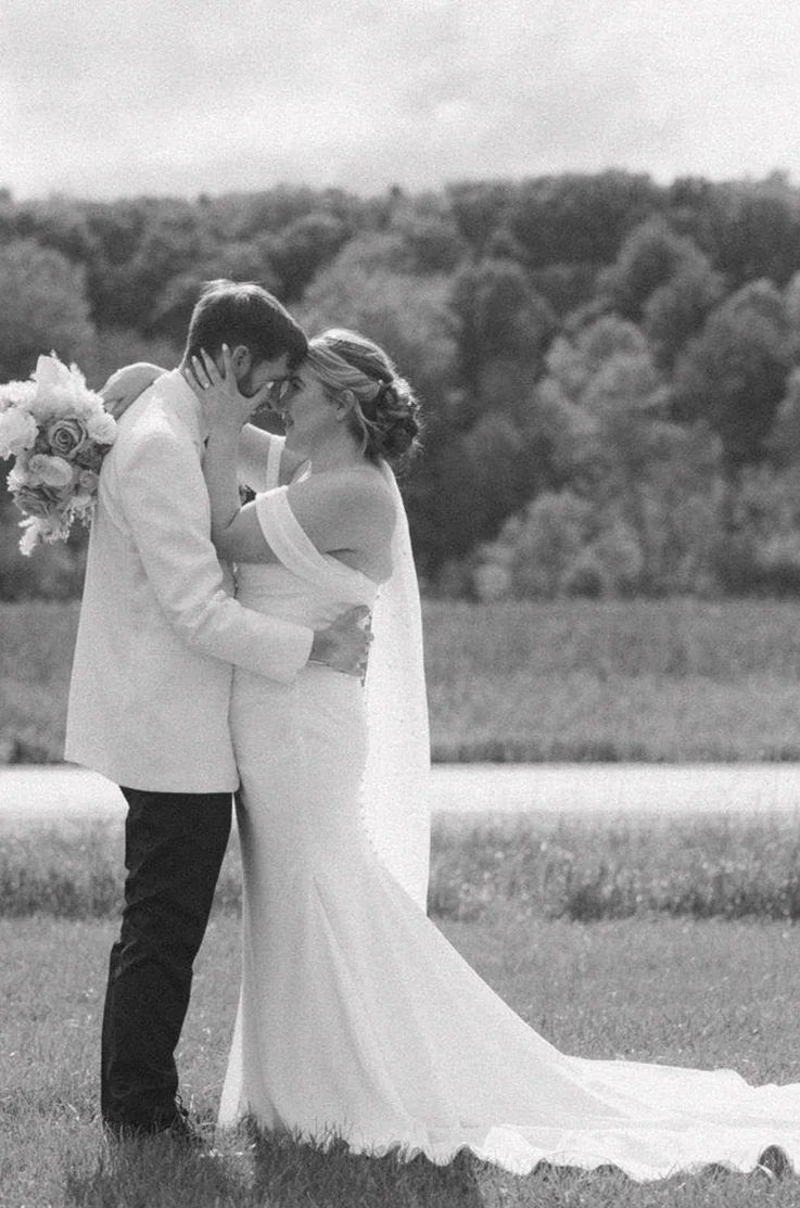 Groom holds his bride in an embrace in a draped crepe bodice gown with sleeves and a veil, by trees in black and white.