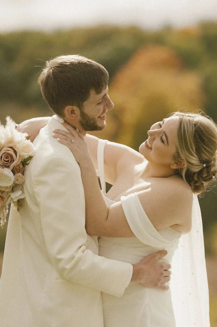Close-up of bride in a crepe off-shoulder gown with a veil and is holding a bouquet while smiling at her groom. 