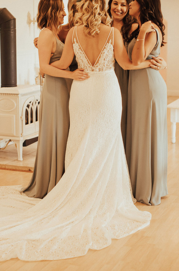 A bride in a lace wedding dress huddles in a circle with her bridesmaids in room with a wood stove, while they all smile.