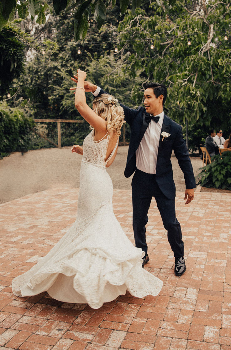 A groom in a suit twirling a bride in a lace wedding dress on a brick patio outside, surrounded by trees.