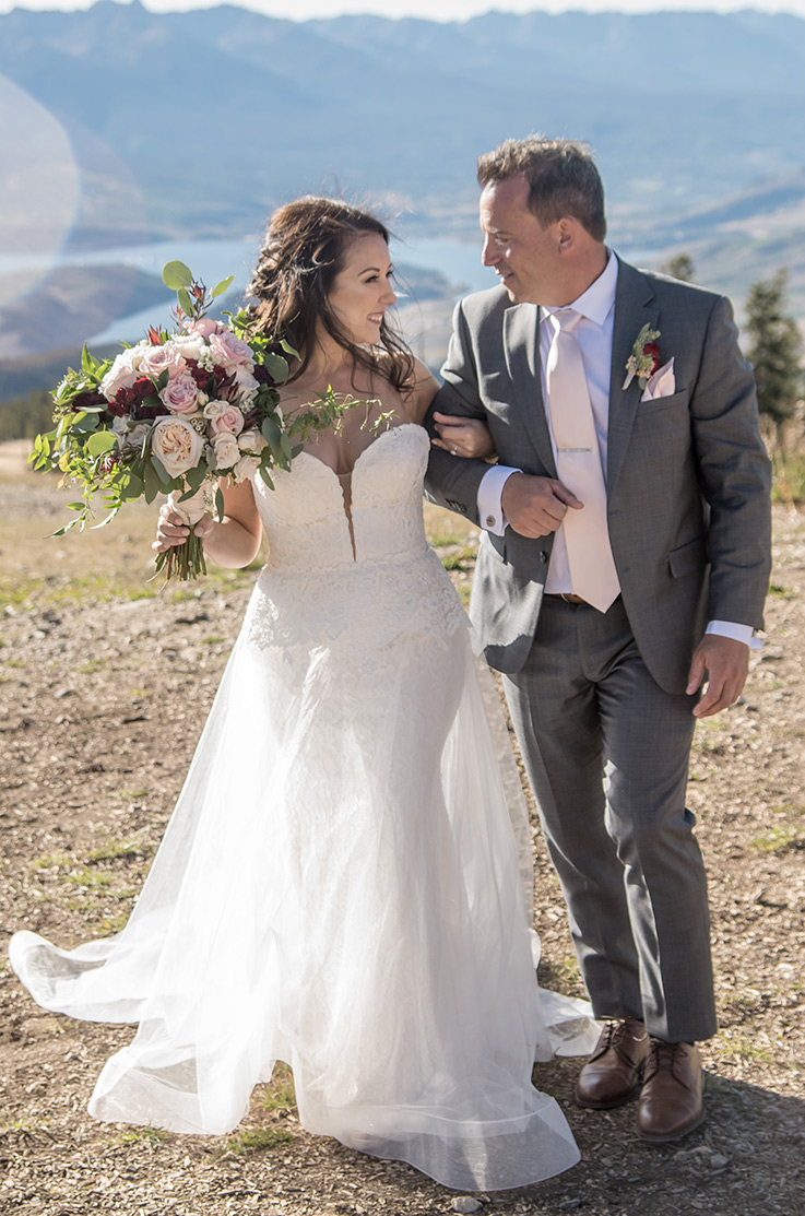 A bride in a strapless lace and tulle wedding dress holds a bouquet and walks with a groom in a suit in a clearing in the mountains.