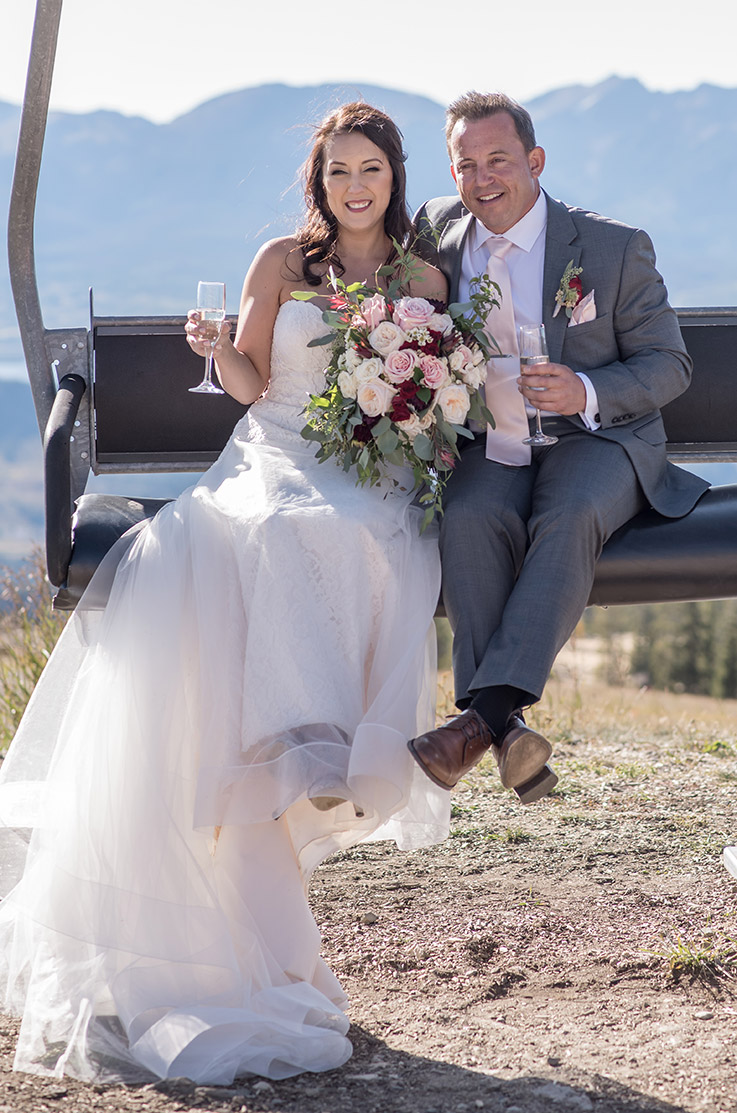 A bride in a lace and tulle wedding dress holding a bouquet and a champagne glass, and a groom in a suit holding a champagne glass, smile and sit on a ski lift with mountains in the background.