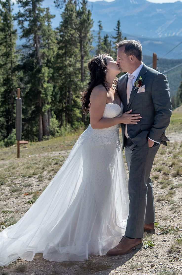 A bride in a lace and tulle wedding dress and a groom in a suit kiss while standing outdoors, with a forest and mountains behind them.