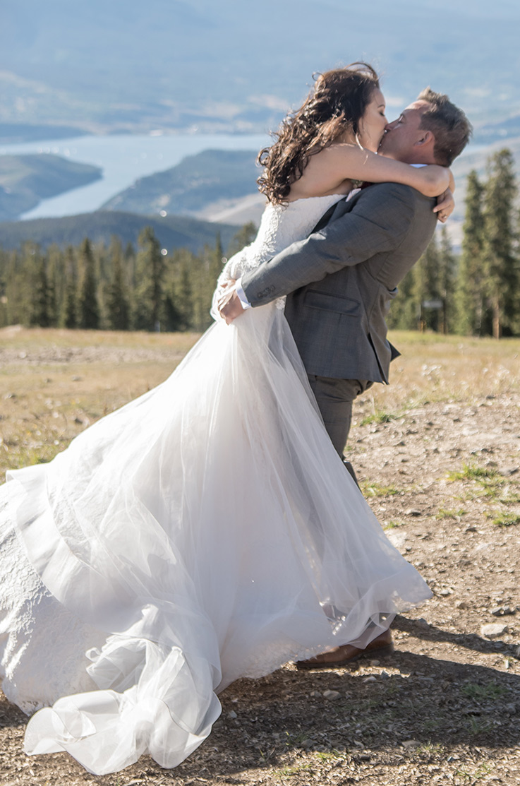 A groom in a suit picks up a bride in a lace and tulle wedding dress as they kiss in a clearing in the mountains.