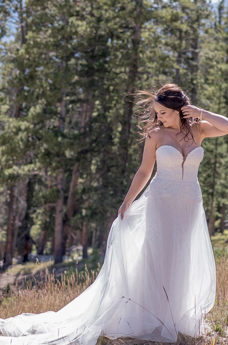 A bride in a strapless lace and tulle wedding dress looks down and to the side while holding her tulle skirt, outside in a forest.