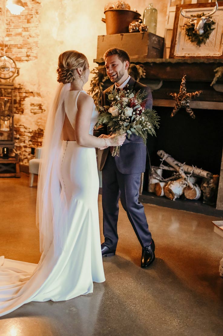 A bride in a keyhole-back wedding dress holds a bouquet and faces a groom in a suit while he smiles, with a brick wall and a fireplace in the background.