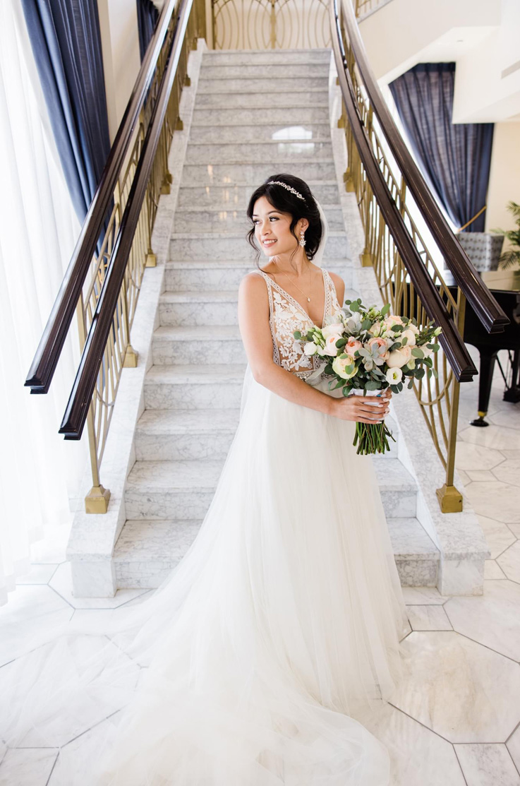 A bride in a lace and tulle wedding dress and a head piece holds a bouquet and looks off to the side while standing in front of a marble staircase indoors.