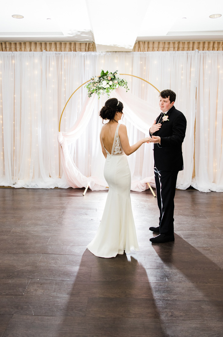 A bride in a fitted wedding dress and a head piece faces a groom in a suit while holding his hand and looking at him, both standing in front of a curtained wall and a circular ceremony decoration.