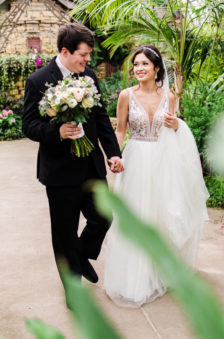 A bride in a lace and tulle wedding dress, a headpiece, and a veil, and a groom in a suit holding a bouquet, walk hand in hand down a path in a garden.