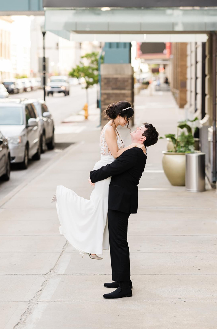 A groom in a suit lifts up a bride in a fitted wedding dress, both of them looking at each other, while standing on a sidewalk in a city.