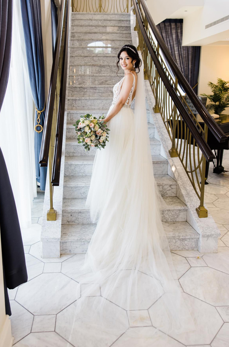 A bride in a lace and tulle wedding dress, a head piece, and a veil, holds a bouquet and smiles while standing on a marble staircase.