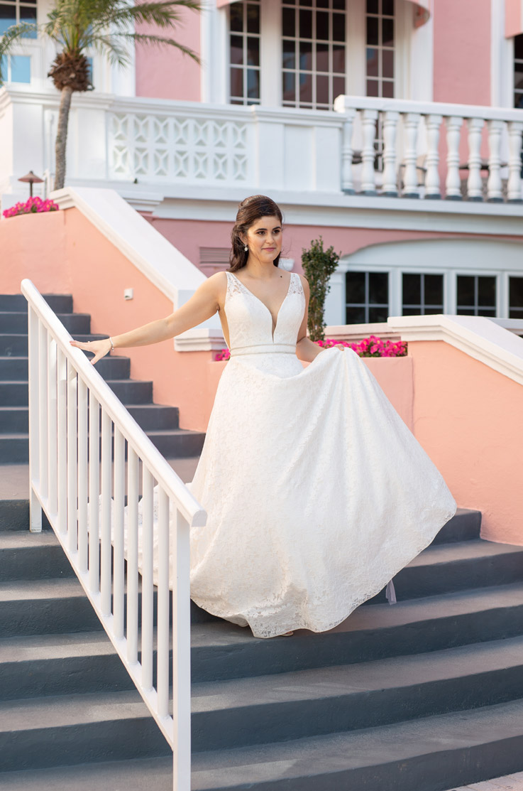 A bride in a sleeveless lace wedding dress walks down a brightly coloured staircase with one hand on the rail and the other holding her skirt up.