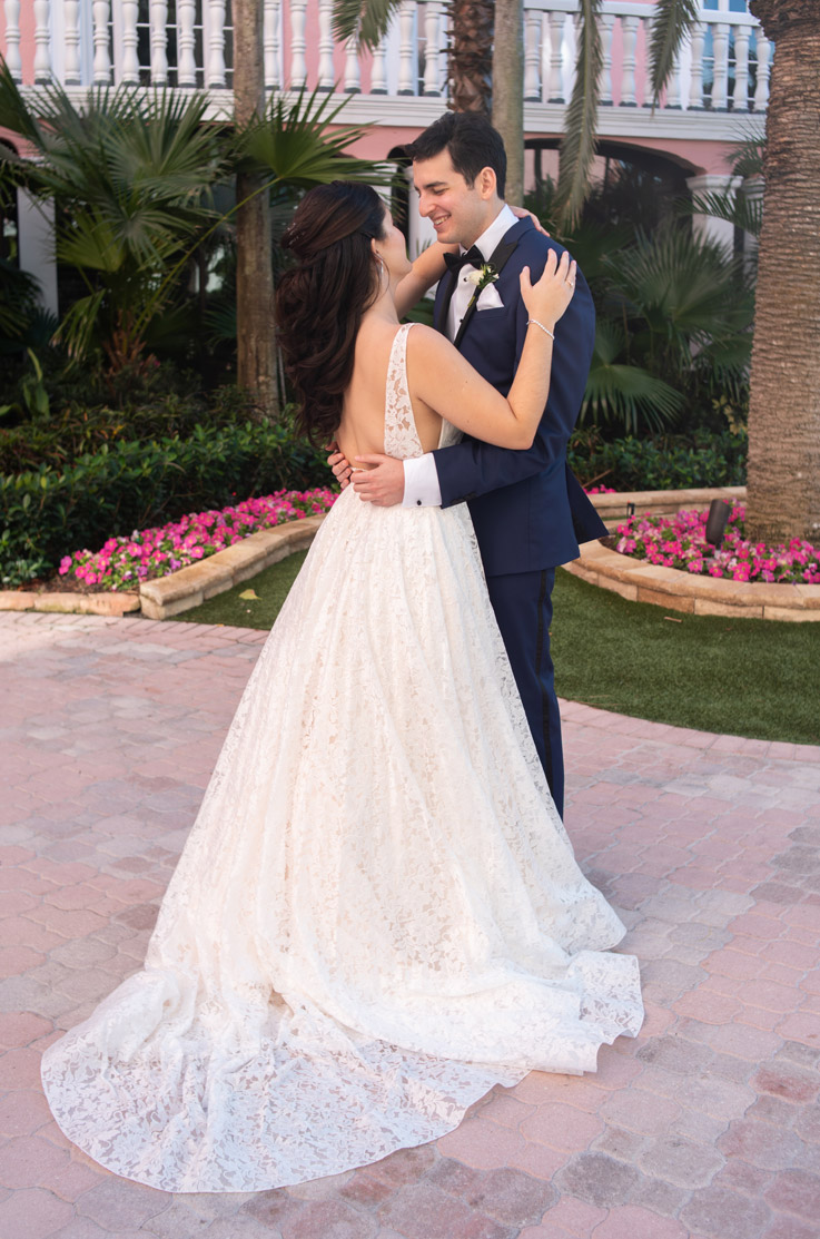 A bride in a lace wedding dress and a groom in a suit hold each other close while standing on a path in a garden outside, with a building in the background.