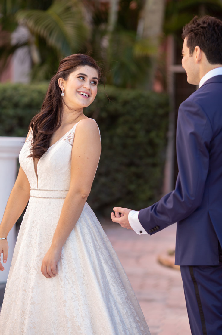 A bride in a sleeveless lace wedding dress smiles and looks over her shoulder at a groom in a suit, while standing outside in a garden.