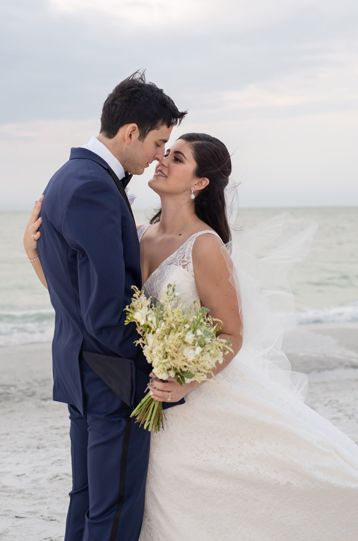 A bride in a lace wedding dress holding a bouquet, and a groom in a suit stand on a sandy beach, holding each other close.