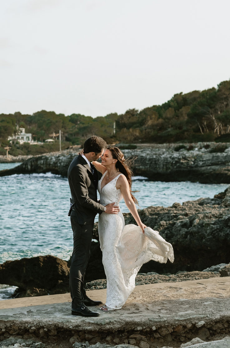 A bride in a lace wedding dress and a groom in a suit stand outside on the rocks beside the ocean, smiling and looking at each other. 
