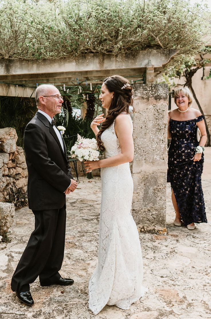 A bride in a lace wedding dress holds a bouquet while smiling and talking to a man in a suit, a woman in a lace dress smiling and looking at them. They are outdoors in front of a stone and wood pergola.