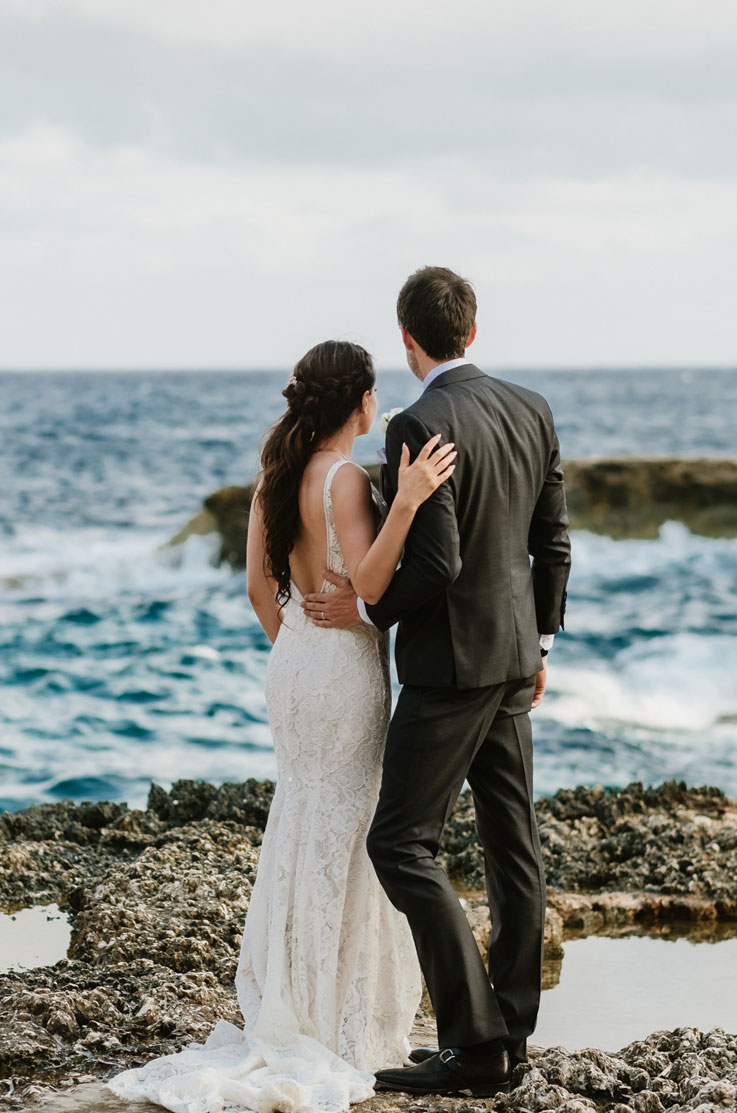 A bride in a lace wedding dress and a groom in a suit stand beside each other on a rocky beach, looking out at the waves of the ocean.