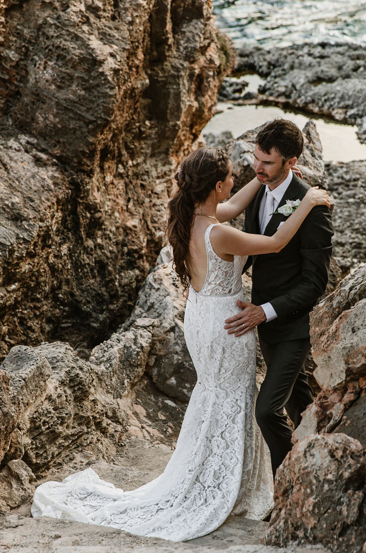 A bride in a lace wedding dress and a groom in a suit stand looking at and holding each other, outside on a rocky path.