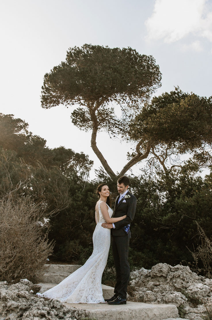 A bride in a lace wedding dress and a groom in a suit stand outside holding each other on a rocky staircase, with trees in the background.