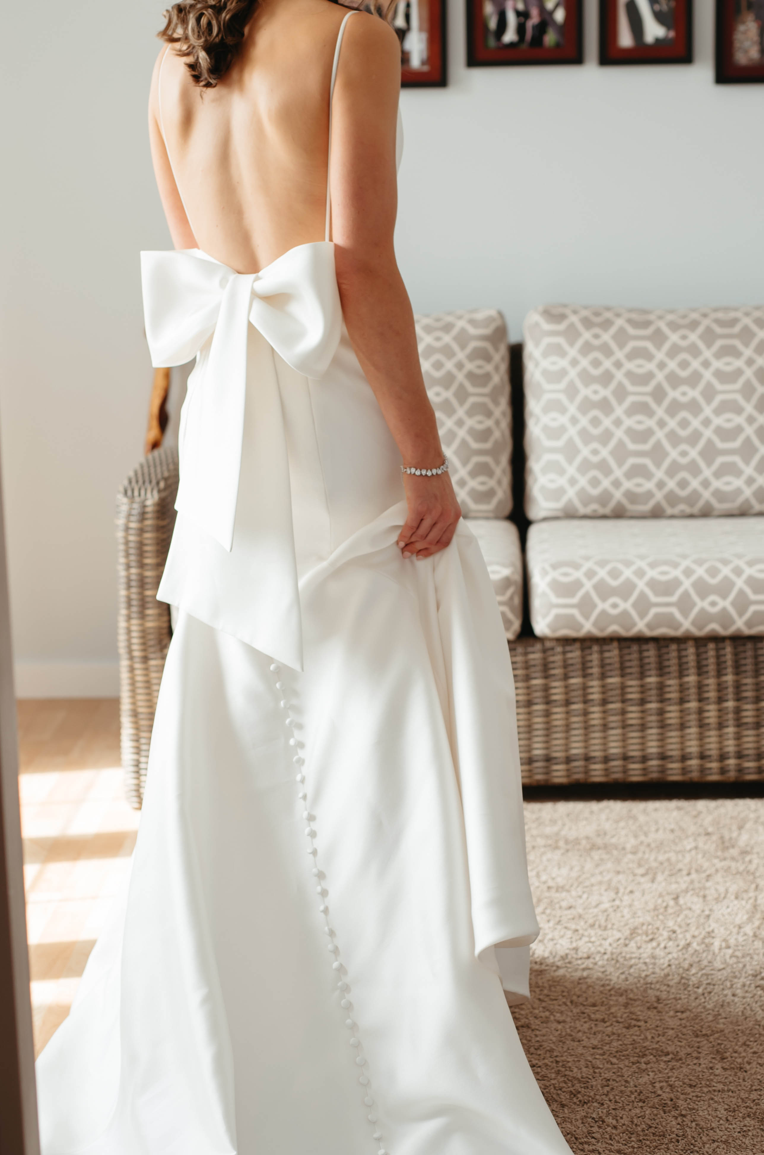 A bride in a wedding dress with a bow on the back stands indoors in front of a couch and family photos on the wall.
