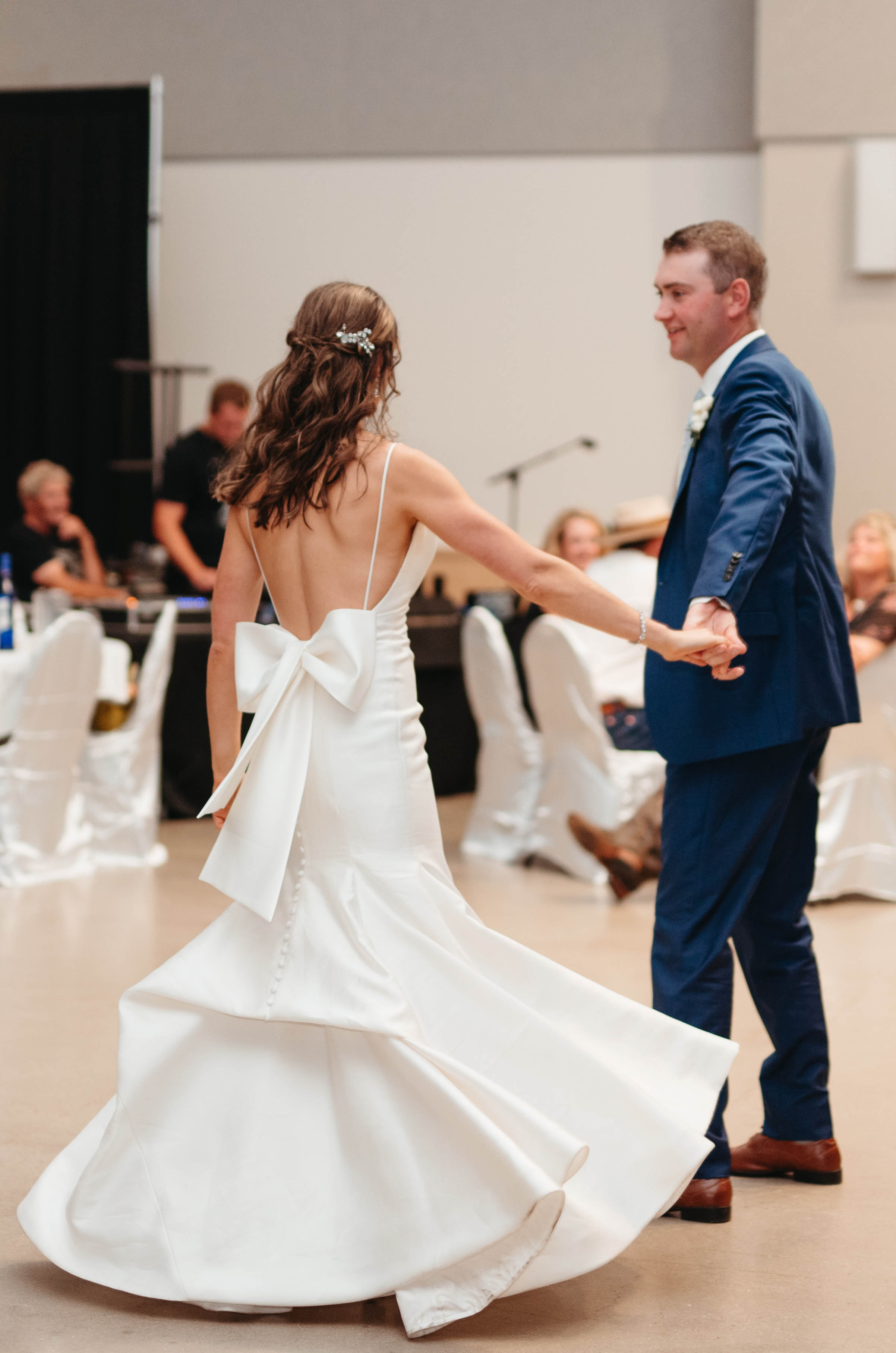 A bride in a wedding dress with a bow on the back and a groom in a suit dance in an event hall, with wedding guests in the background.
