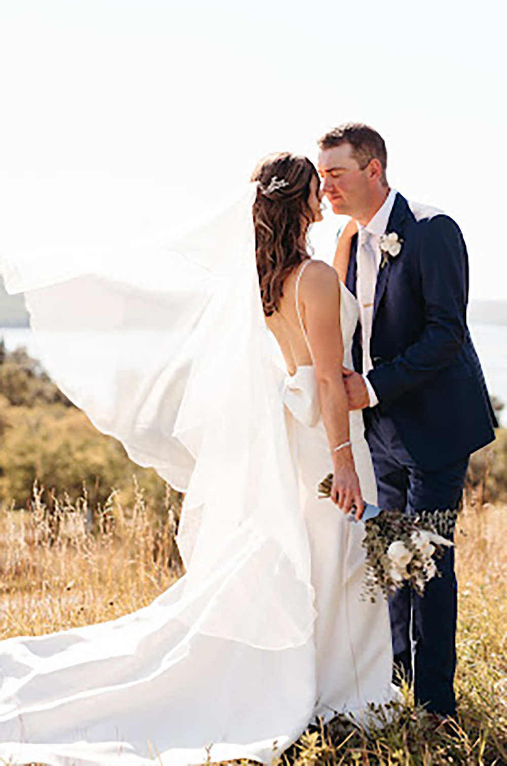 A bride in a fitted wedding dress and veil, and a groom in a suit face each other and hold each other close while standing outdoors in a grassy field.