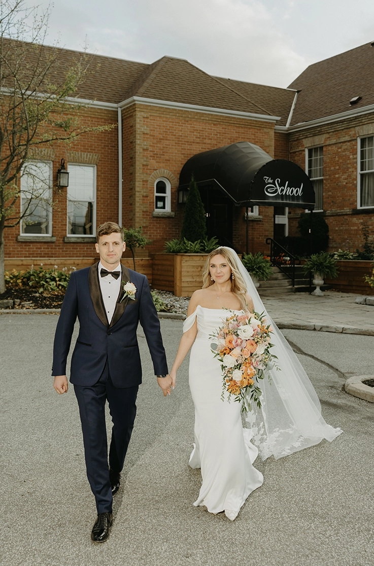 Bride in an off-shoulder charmeuse satin wedding dress with sleeves and a veil, walks next to groom and holds a large bouquet.
