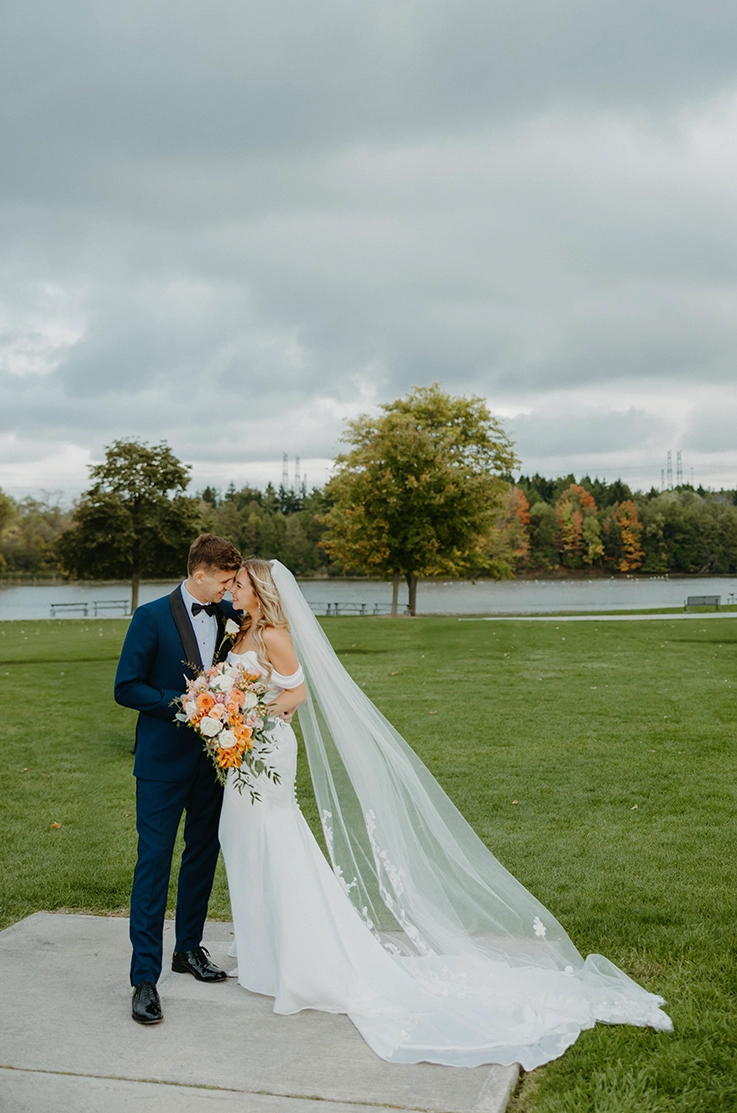 Bride in a charmeuse satin fit and flare gown with draped sleeves and a veil, poses with her groom by a lakefront.