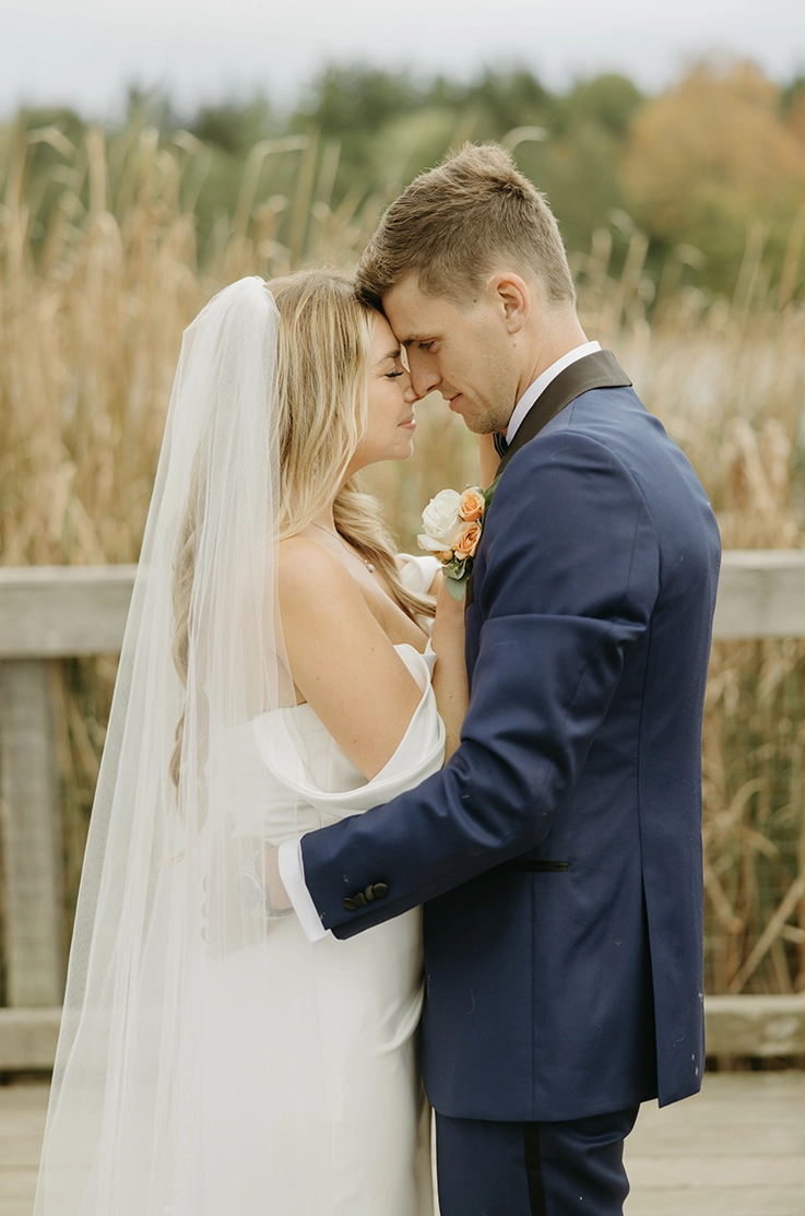 Close-up of groom outdoors embracing bride in a charmeuse satin gown with draped off-shoulder sleeves and a veil by a field.