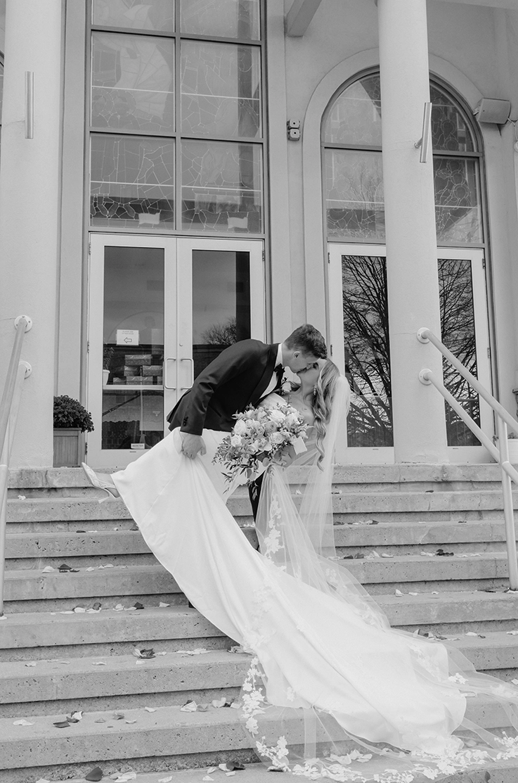 Groom dips his bride in a charmeuse satin fitted gown with a flowing train and lace-trim veil, on steps in front of a building, in black and white.