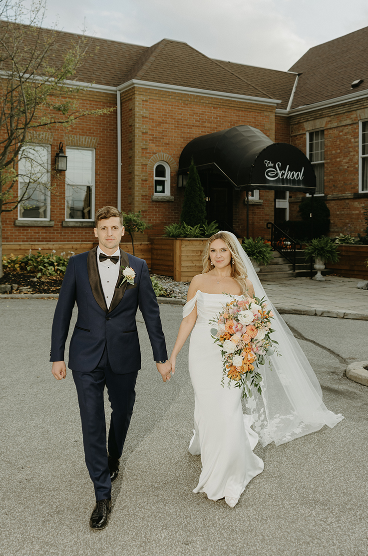 Bride in an off-shoulder charmeuse satin wedding dress with sleeves and a veil, walks next to groom and holds a large bouquet.