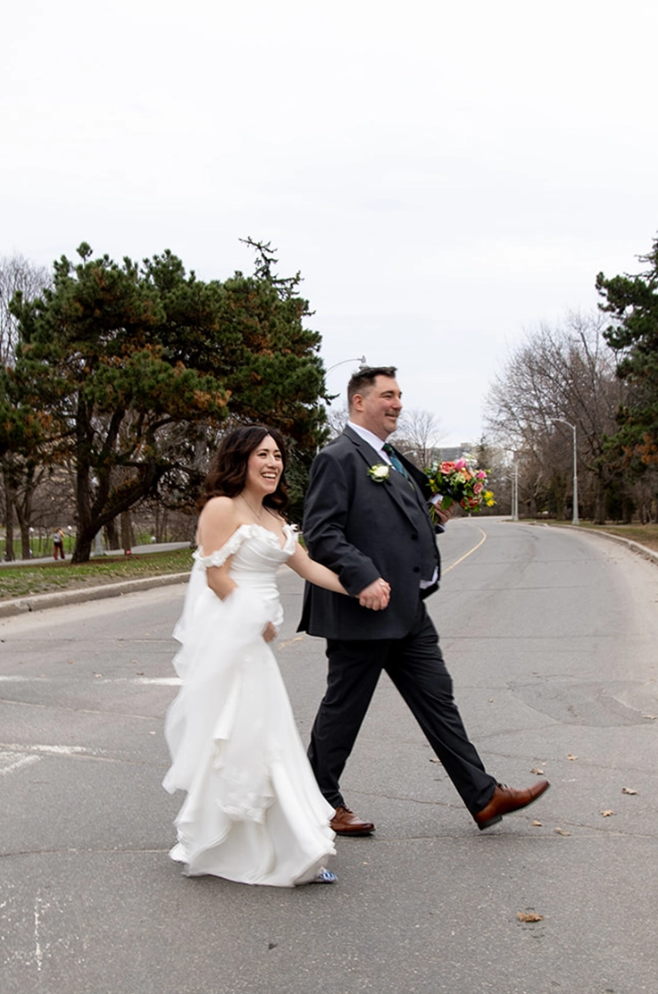 Bride in an off-shoulder fit and flare gown with draped sleeves, holds her train up while walking beside groom along a street.