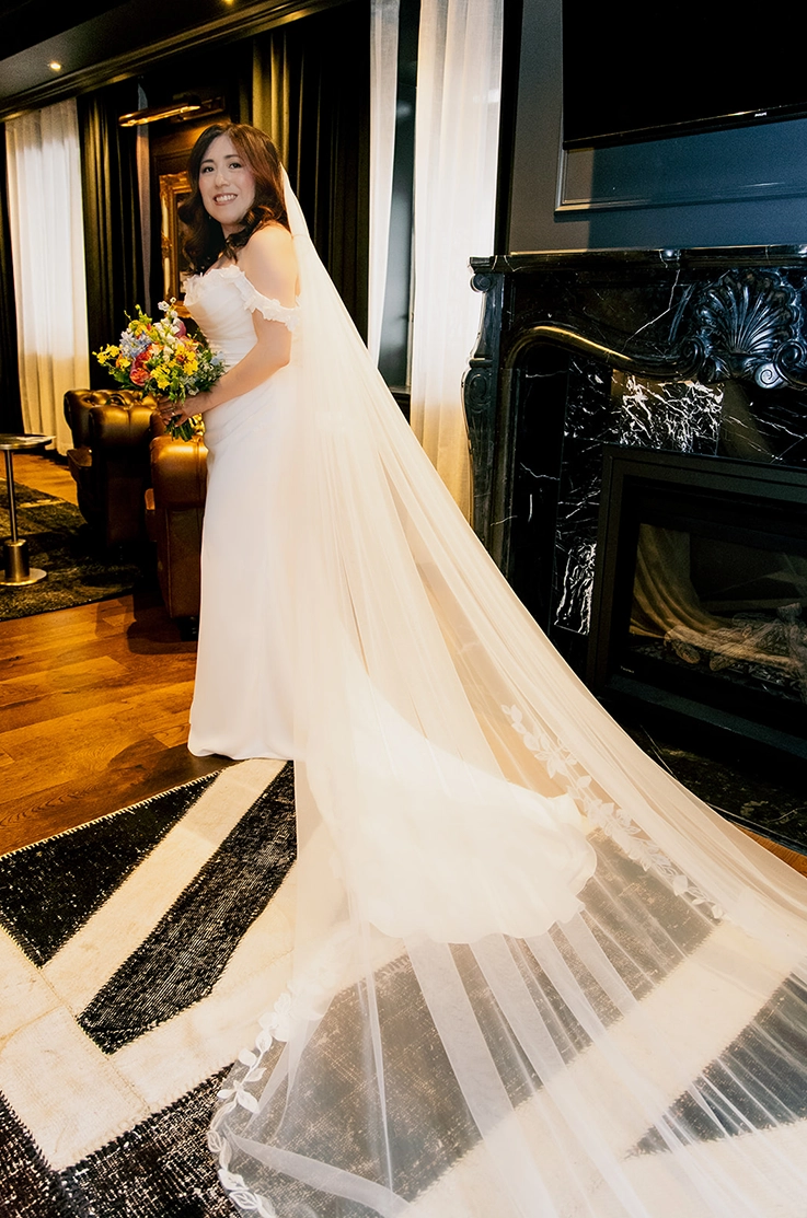 Bride smiles in a strapless charmeuse satin wedding dress with lace detail and a veil, holding a bouquet and poses indoors.