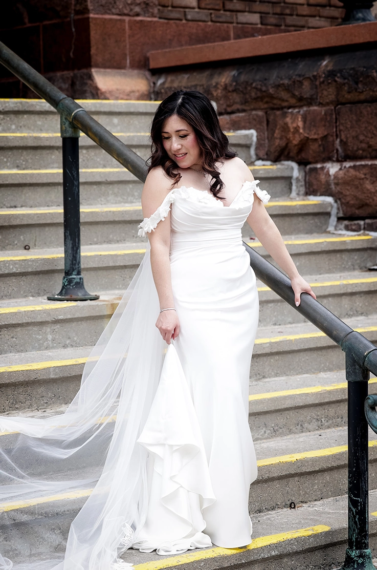 Bride poses on steps outside a brick building, in a charmeuse satin gown with a draped bodice, lace and a veil by a railing.