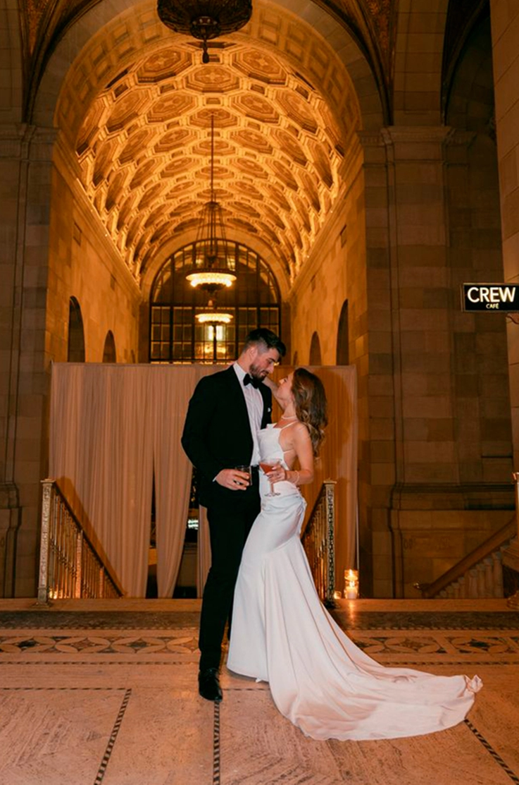 Bride in a sleek cowl neck satin gown, is holding a drink and faces groom at the top of stairs in an elegant interior.