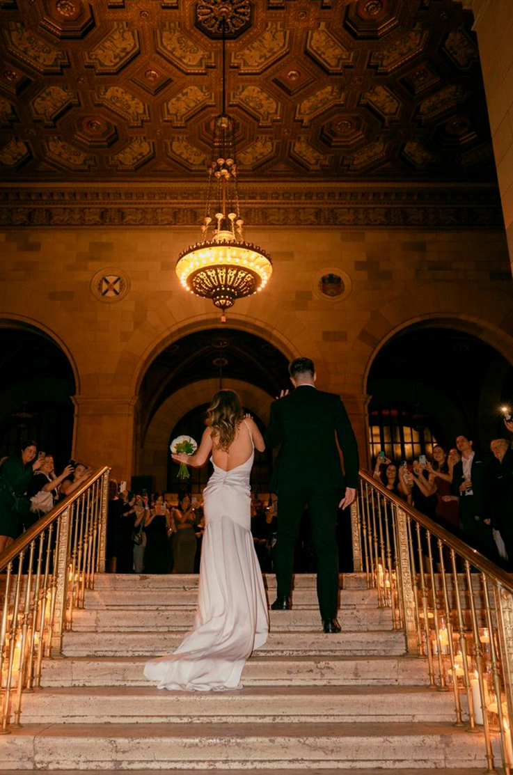 Bride walks up a grand staircase lined with candles in a charmeuse satin fitted gown with a low back beside her groom.
