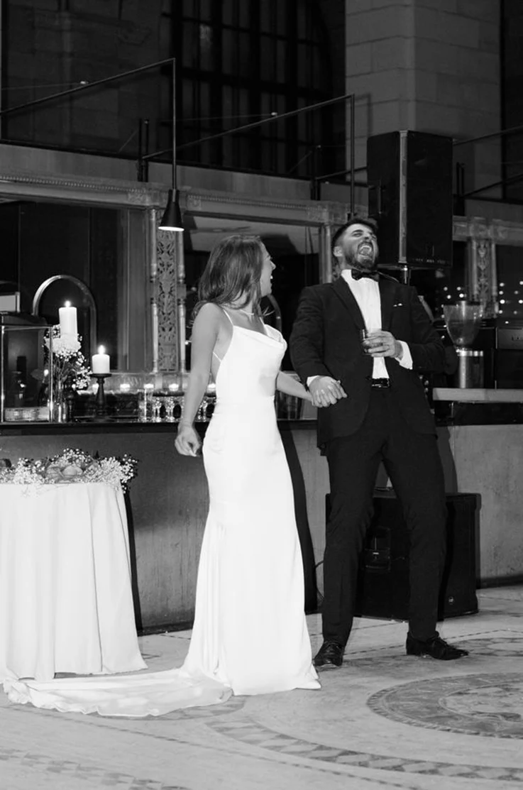 Bride in a charmeuse satin fit and flare wedding dress with a cowl neck, laughs beside groom at the ceremony.