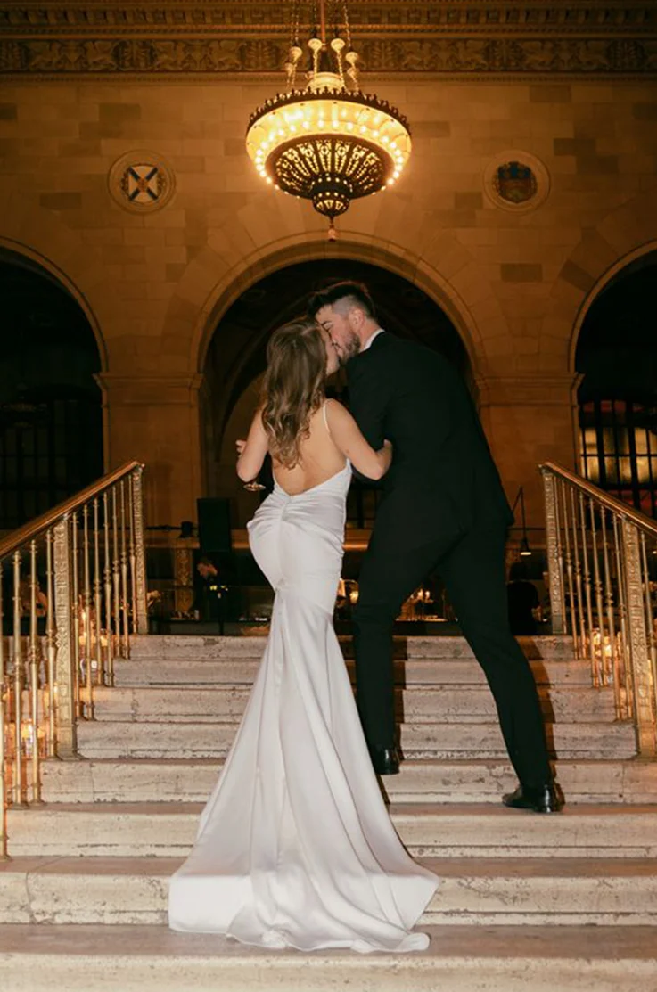 Bride in a low back charmeuse satin gown with thin straps and draping, kisses her groom on a candlelit staircase. 