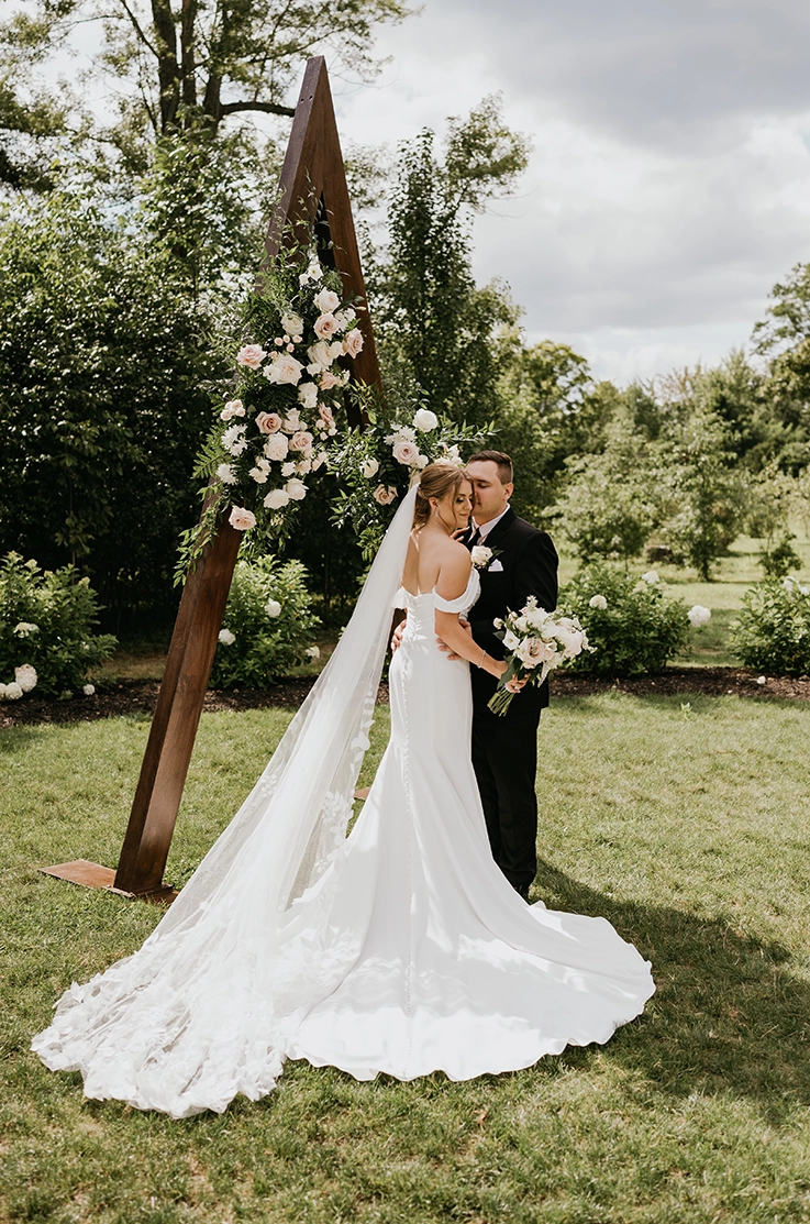 Groom embraces bride in an off shoulder fit and flare crepe gown with a veil, holding a bouquet at a ceremony arch.