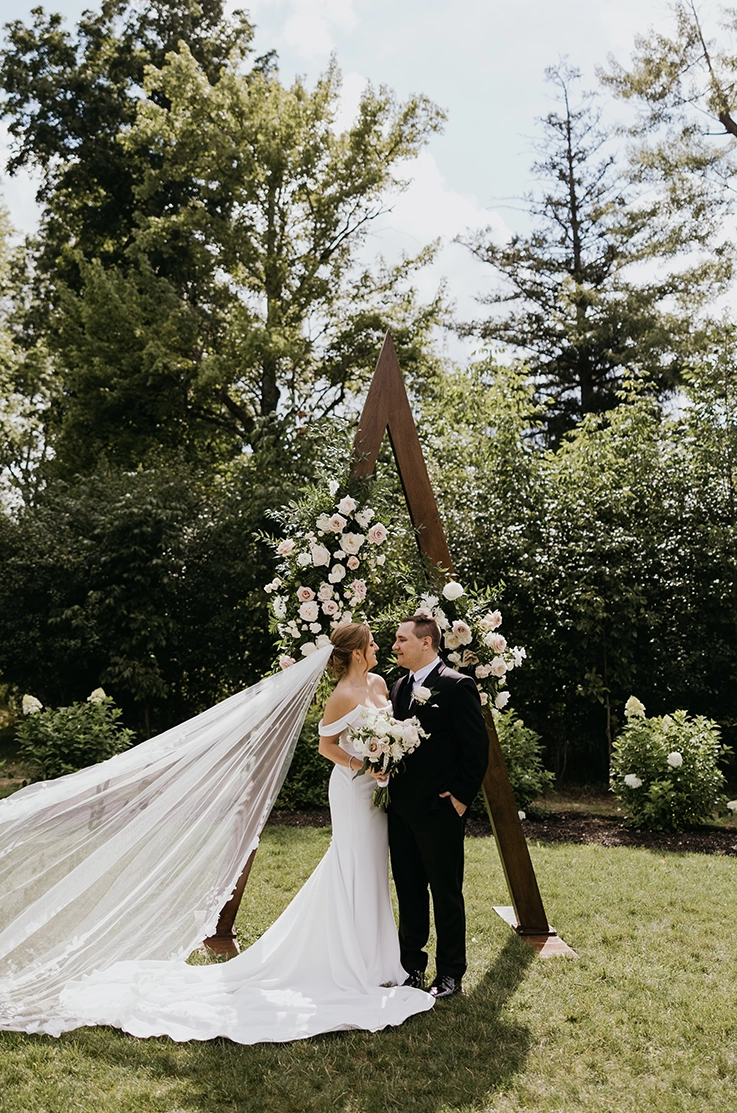 Bride standing in an off-shoulder fitted crepe wedding dress with a long veil, holding a bouquet beside the groom outdoors.