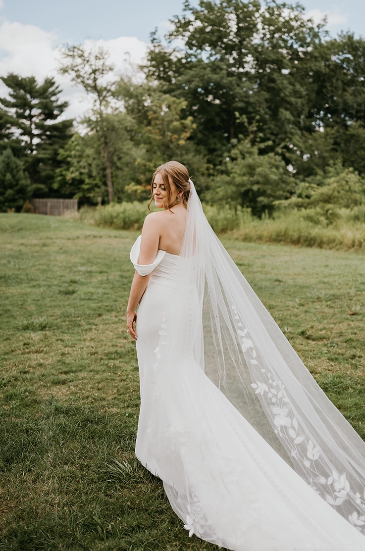 Bride glances over her shoulder in an open meadow, wearing an off-shoulder gown with a draped bodice, veil and fit and flare skirt.