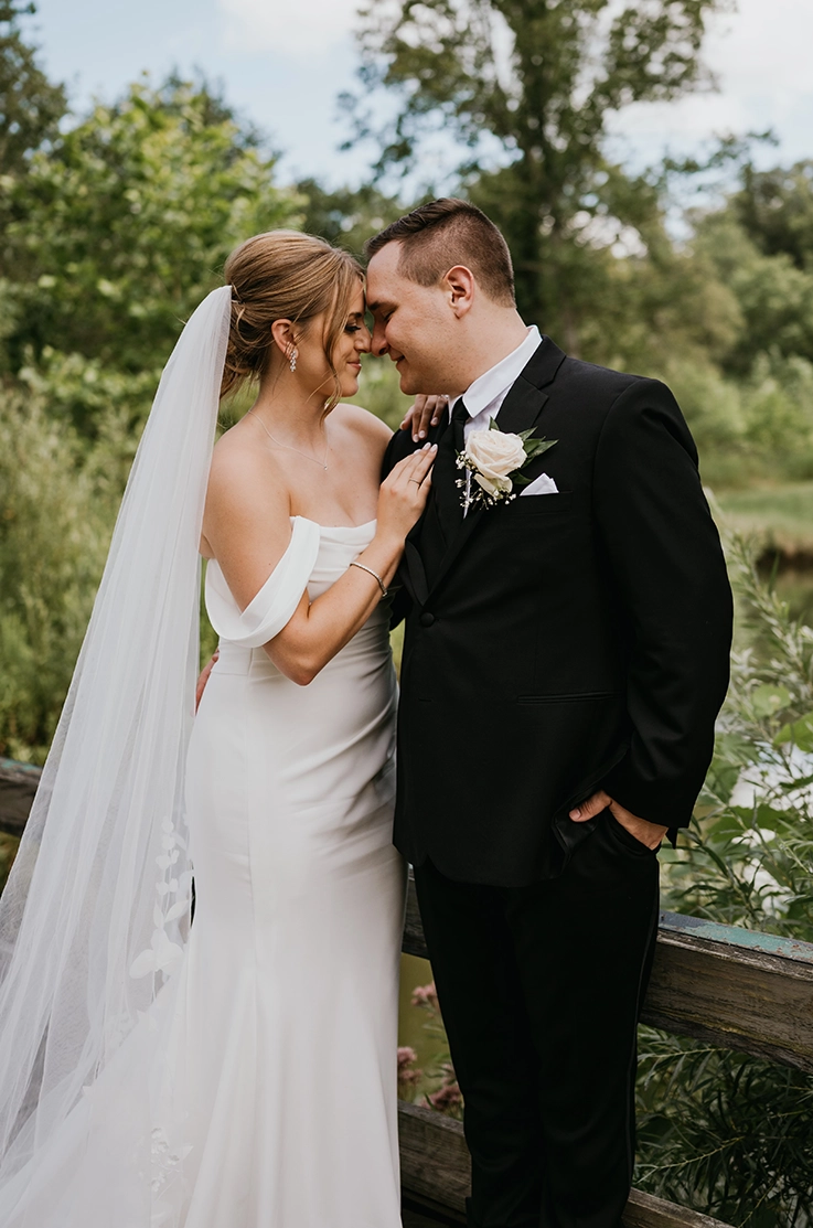 Bride in a draped bodice crepe gown with off-shoulder sleeves and a veil, poses with her groom at a garden bridge. 