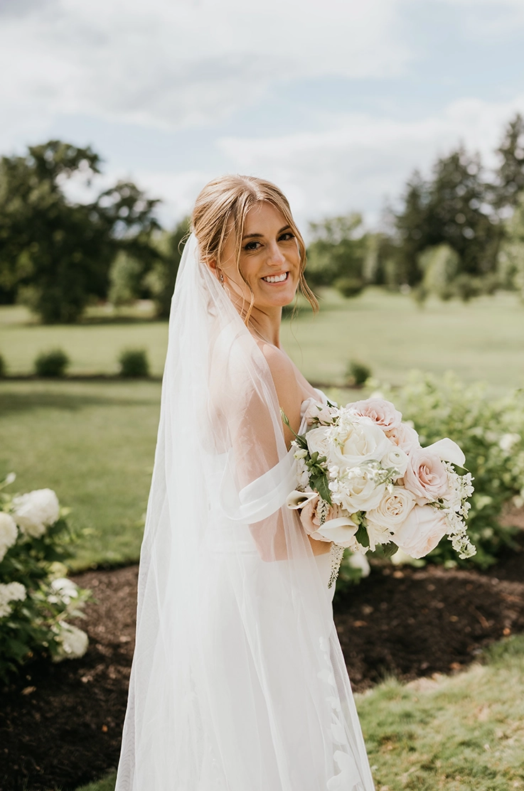Smiling bride in a fit and flare crepe wedding gown with draped sleeves and a veil, holding a blush bouquet outdoors.