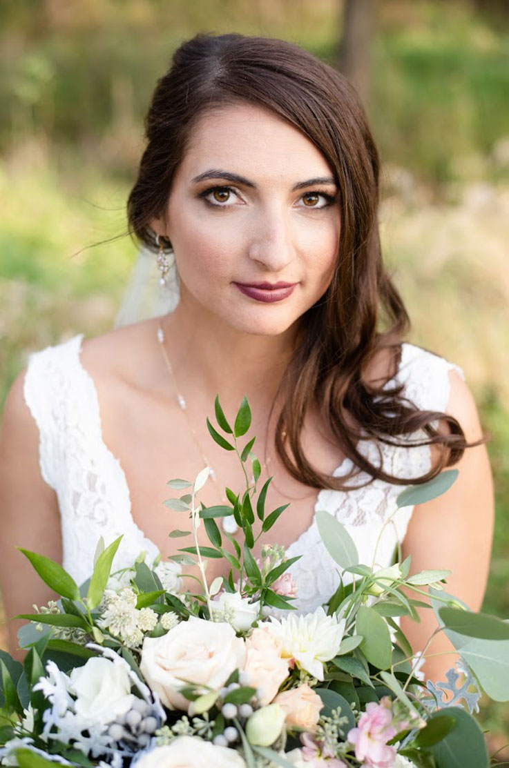 A bride in a lace wedding dress smiles and holds a bouquet while standing outside.