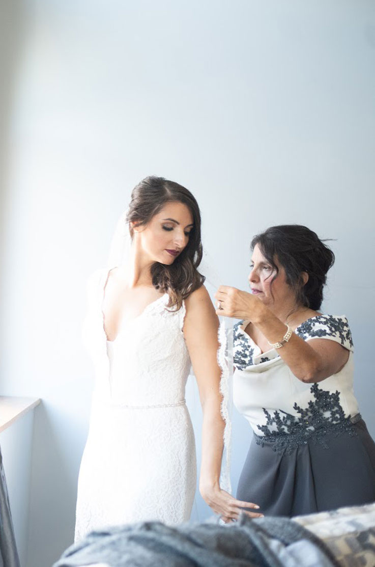A woman in a cap sleeve dress helps a bride in a lace wedding dress with her veil, while standing inside in front of a blank wall.