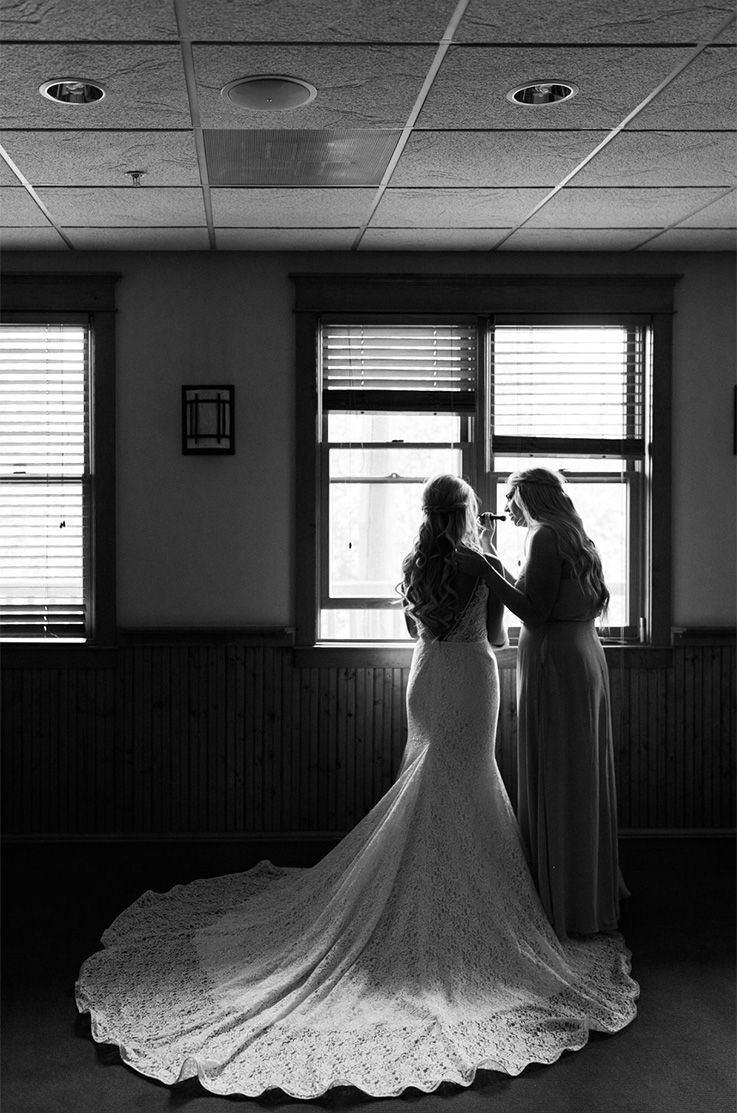 A bride in a lace wedding dress with a long train and a woman in a long dress stand by a set of windows and look out.
